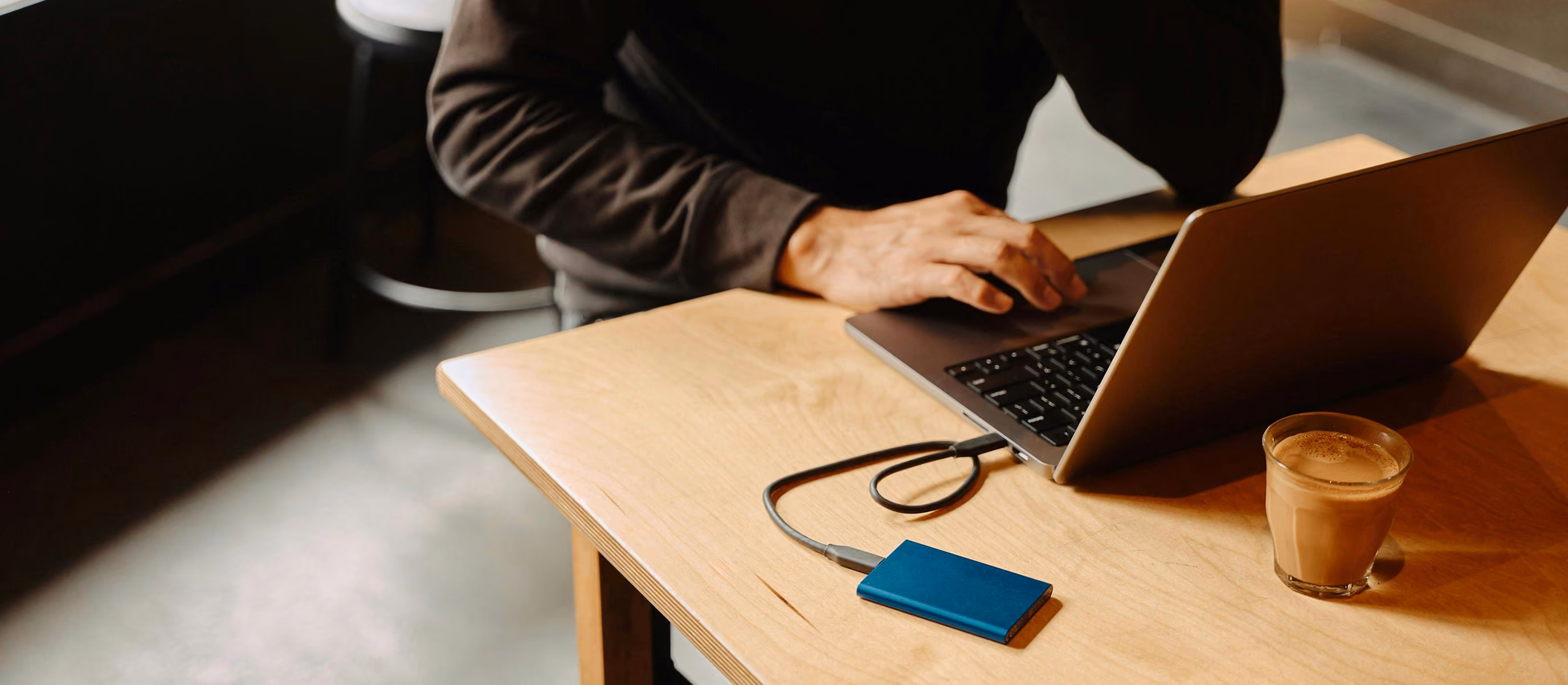Person typing on a laptop connected to a blue Samsung external SSD on a wooden table with a glass of coffee nearby.
