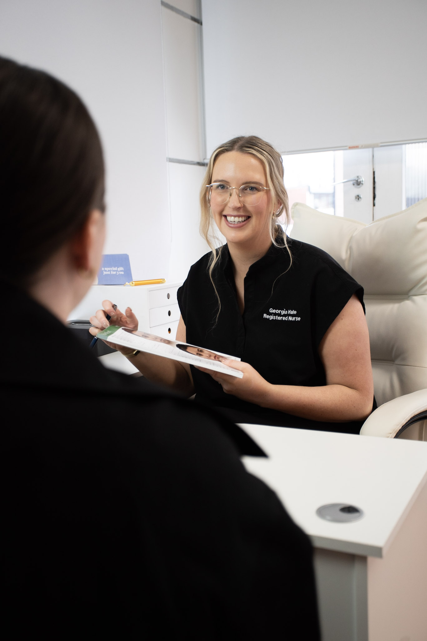 Smiling registered nurse in black uniform holding a brochure and speaking with a patient across the desk.