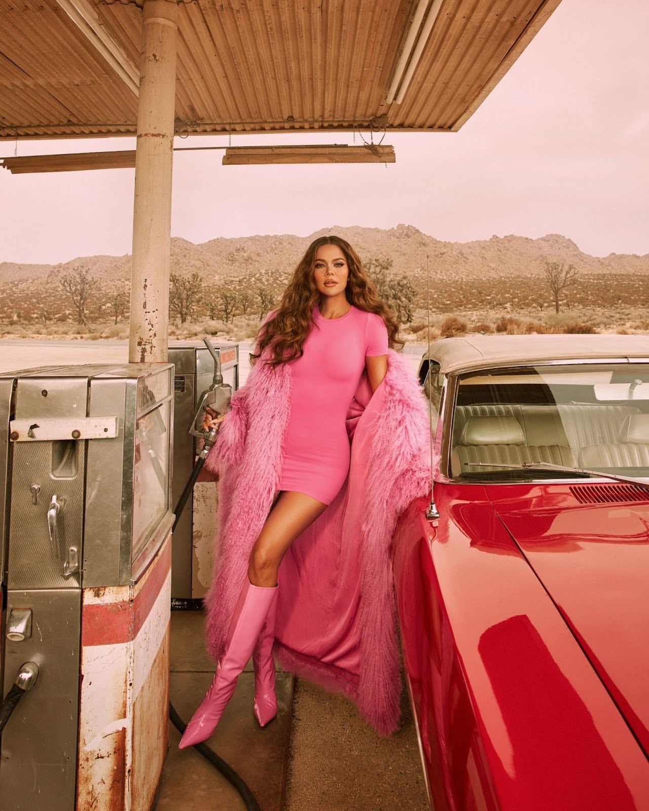 Woman in pink dress, boots, and fur coat fueling a red vintage car at a desert gas station.