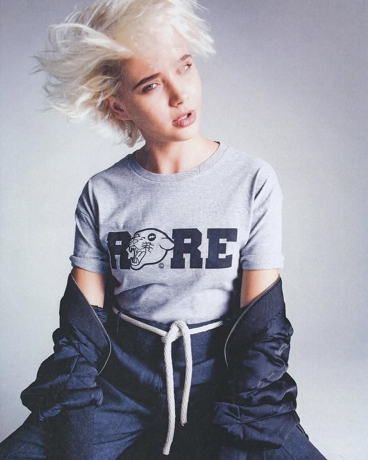 Young person with windswept blonde hair wearing a grey ROAR t-shirt and black jacket.