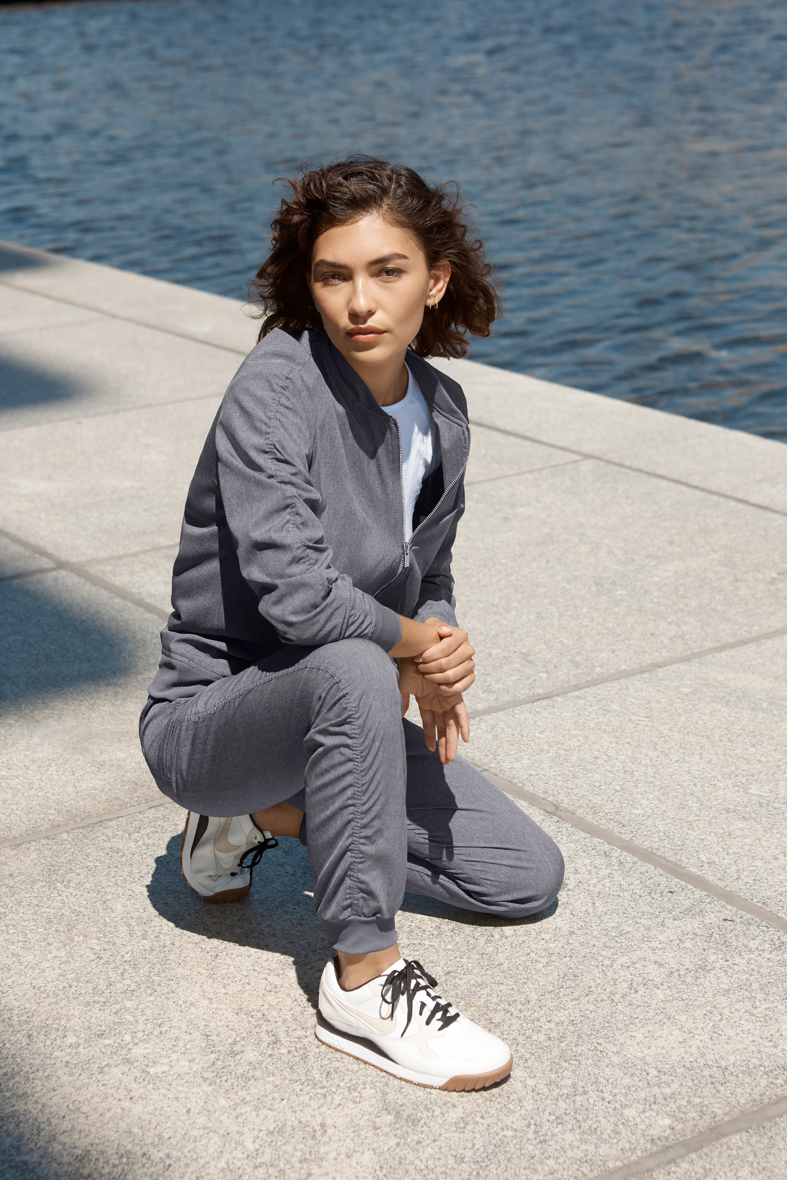 Woman in gray ruched jogger pants and white sneakers stands near water with arms crossed.