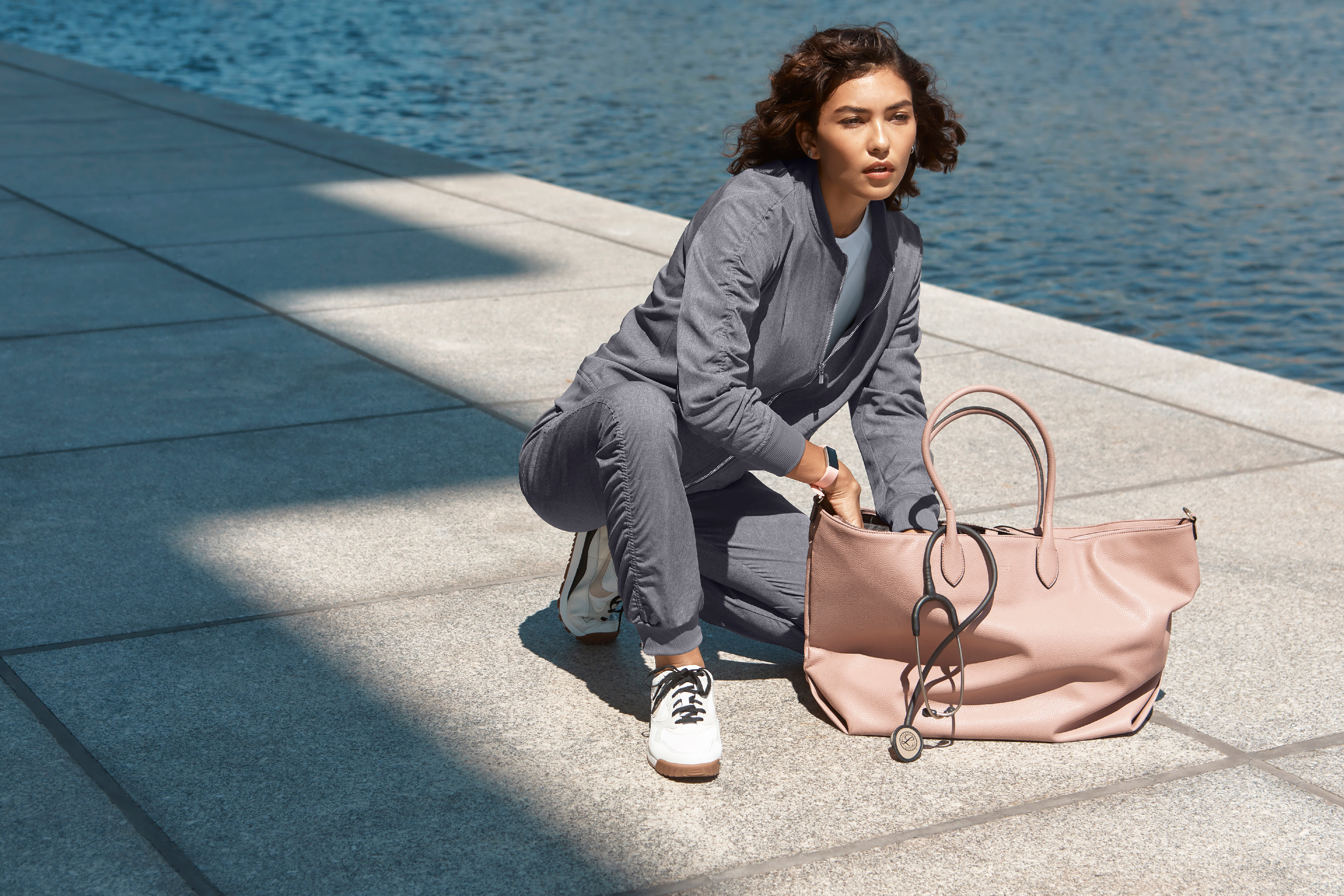 Woman in gray ruched bomber jacket and jogger pants crouching outdoors by water with a pink handbag.