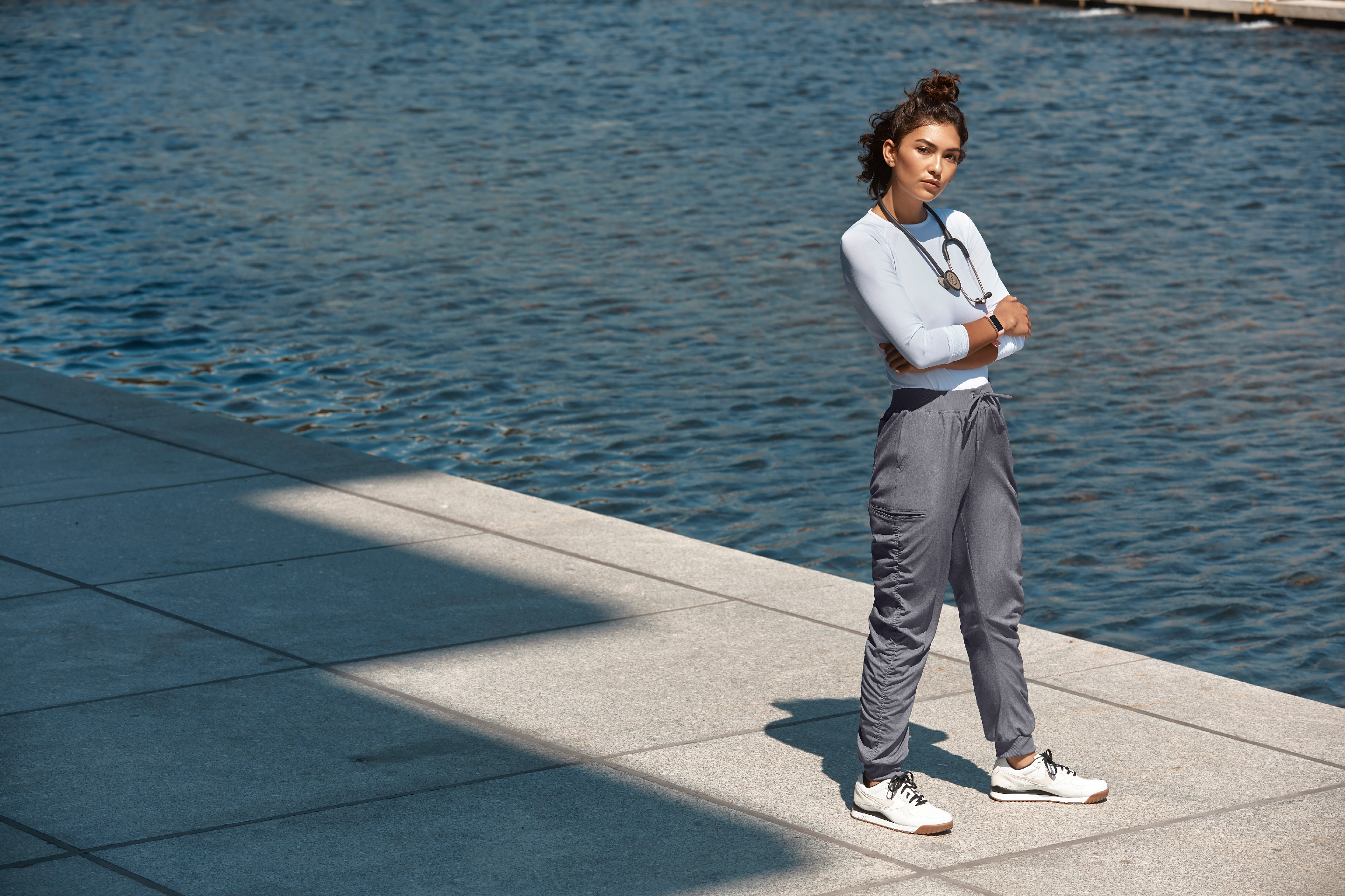 Woman in gray ruched jogger pants and white sneakers stands near water with arms crossed.