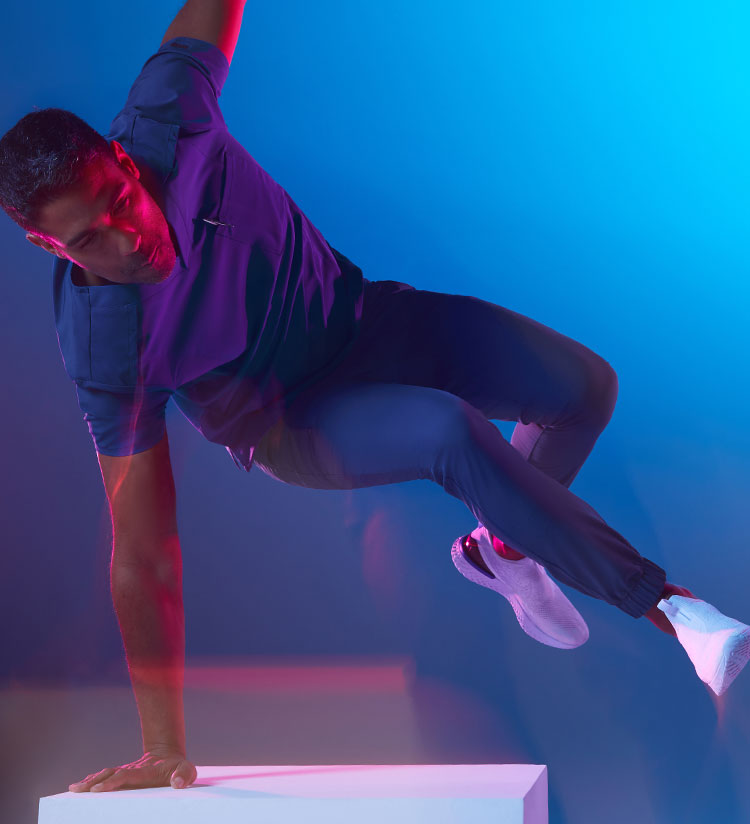 Man in blue scrubs balancing on one hand on a white block against blue light background.