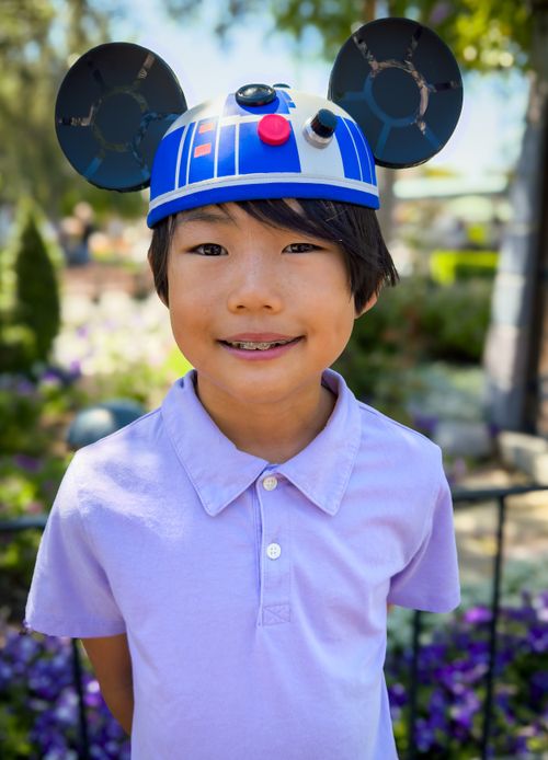 A young boy in a purple shirt stands in a park wearing R2D2 Mickey ears and smiling