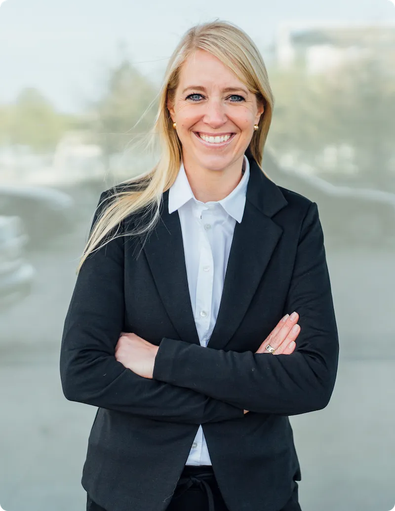 Eine blonde Frau in schwarzem Blazer mit verschränkten Armen lächelt vor einem verschwommenen städtischen Hintergrund.