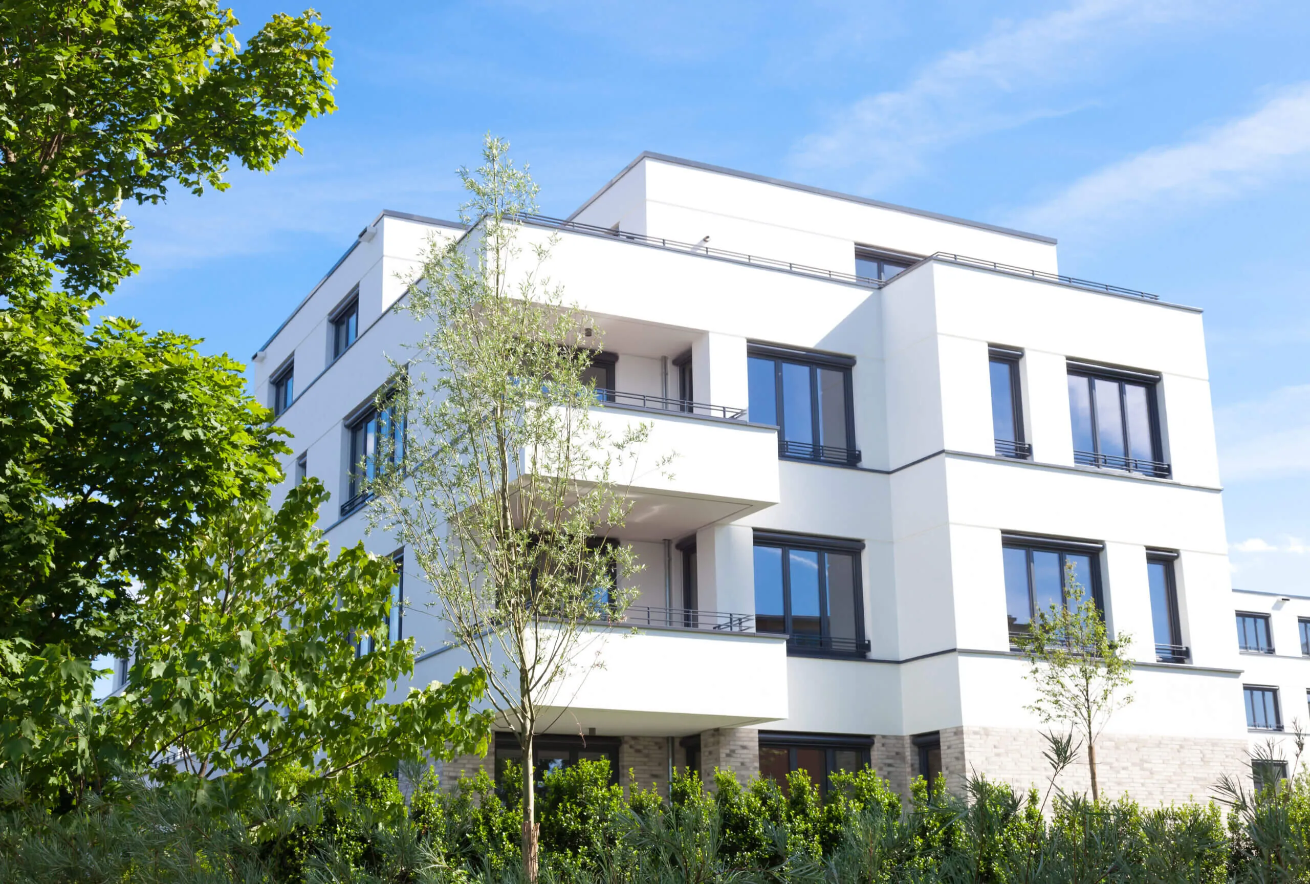 Moderner weißer Wohnblock mit großen Fenstern und Balkonen, umgeben von Bäumen und Sträuchern.