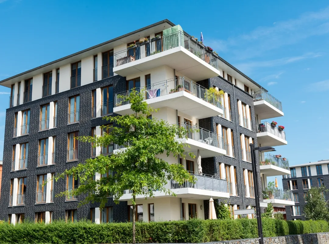 Modernes Mehrfamilienhaus mit Balkonen, begrünten Flächen und einem Baum davor unter blauem Himmel.
