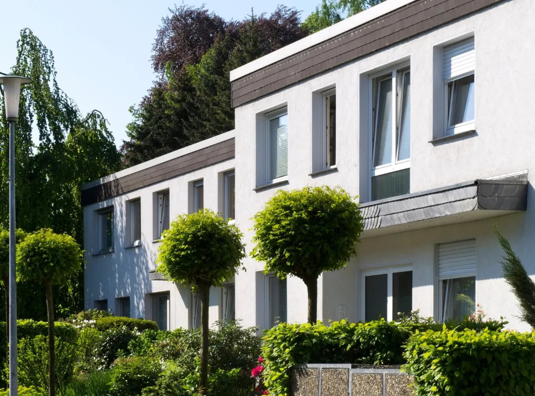Moderne weiße Gebäudefassade mit mehreren Fenstern und gut gepflegtem Garten mit rund geschnittenen Bäumen und grünen Sträuchern.