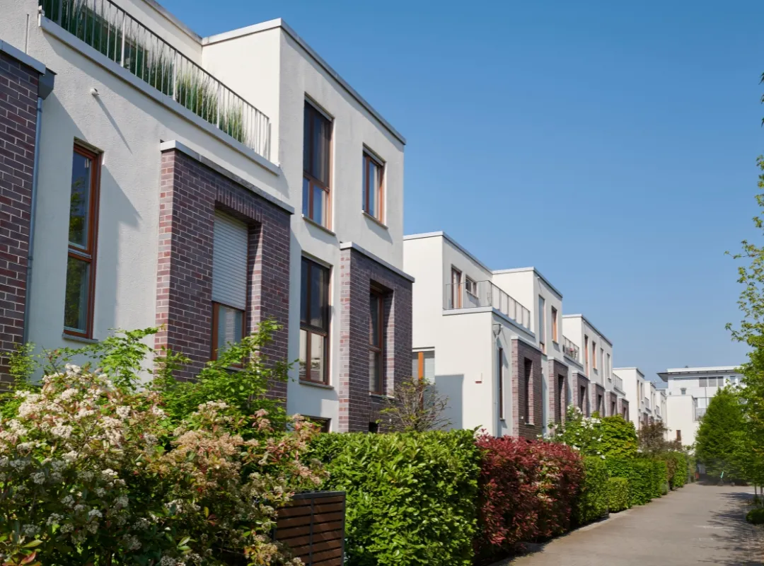 Moderne Reihenhäuser mit weißen Fassaden und Ziegelverkleidung, umgeben von blühenden Sträuchern und Bäumen unter klarem blauem Himmel.