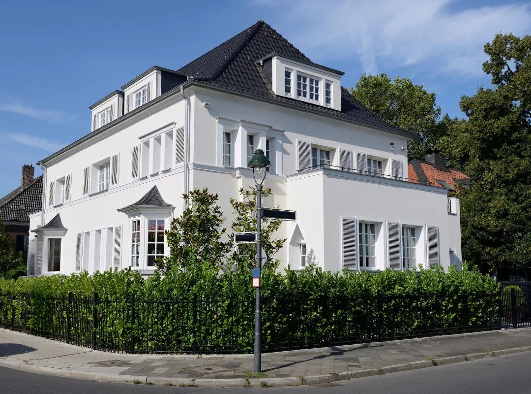 Modernes zweistöckiges weißes Wohnhaus mit schwarzen Dächern an einer Straßenecke, umgeben von grünen Hecken und Bäumen.