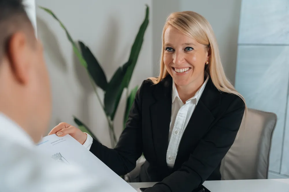 Eine lächelnde blonde Frau in schwarzem Blazer übergibt einem Mann ein Dokument während eines Gesprächs in einem Büro.