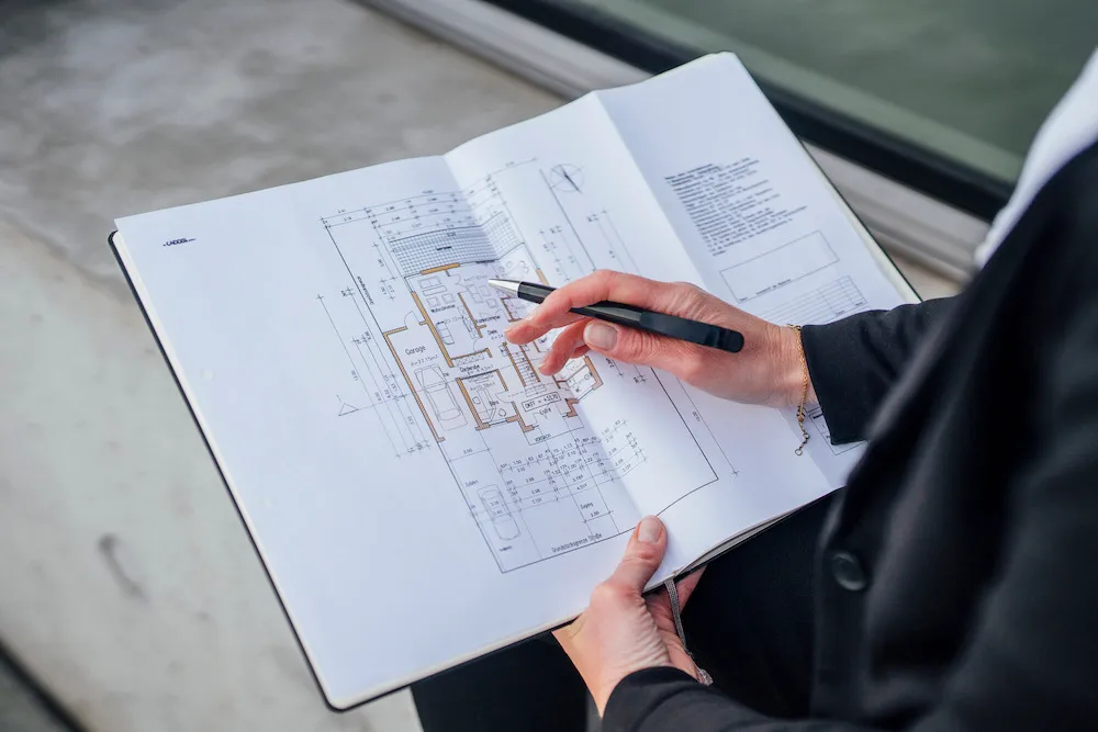 Person hält einen geöffneten Ordner mit einem Architekturplan und zeigt mit einem Stift auf die Zeichnung.