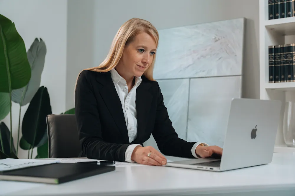Geschäftsfrau mit blondem Haar in schwarzem Blazer arbeitet an einem Laptop an einem weißen Schreibtisch in einem modernen Büro.