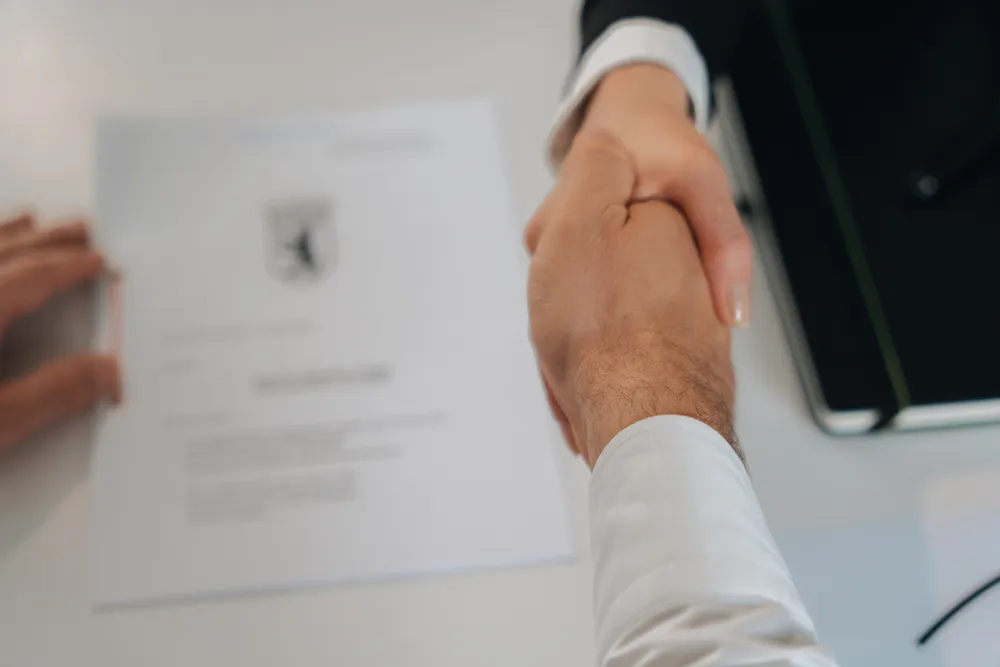 Zwei Personen geben sich die Hand über einem Tisch mit einem Dokument und einem Notizbuch.