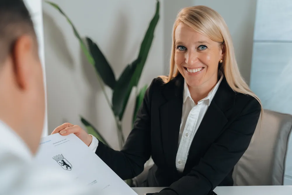 Lächelnde blonde Frau in schwarzem Blazer übergibt einer anderen Person ein Dokument mit einem Wappen.