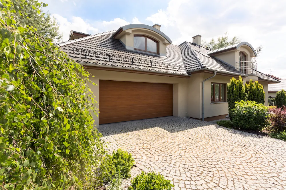 Ein freistehendes Haus mit grauem Ziegeldach, einer großen Holztor-Garage und gepflasterter Auffahrt, umgeben von grünen Pflanzen.