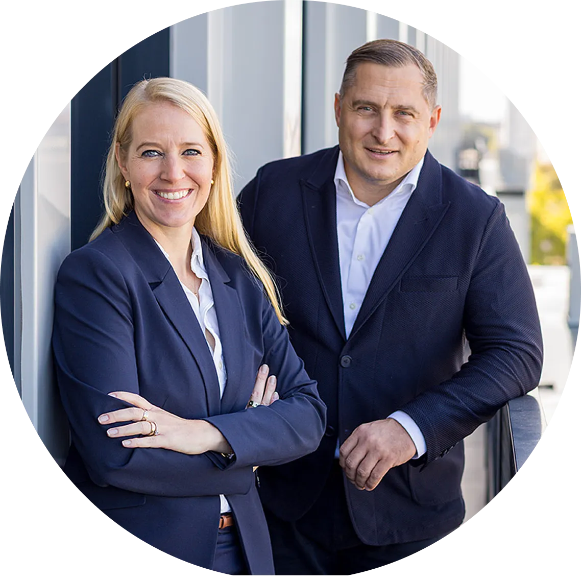 Zwei Geschäftsleute, eine blonde Frau und ein Mann mit dunkelblauem Anzug, lächeln vor einem modernen Gebäude.