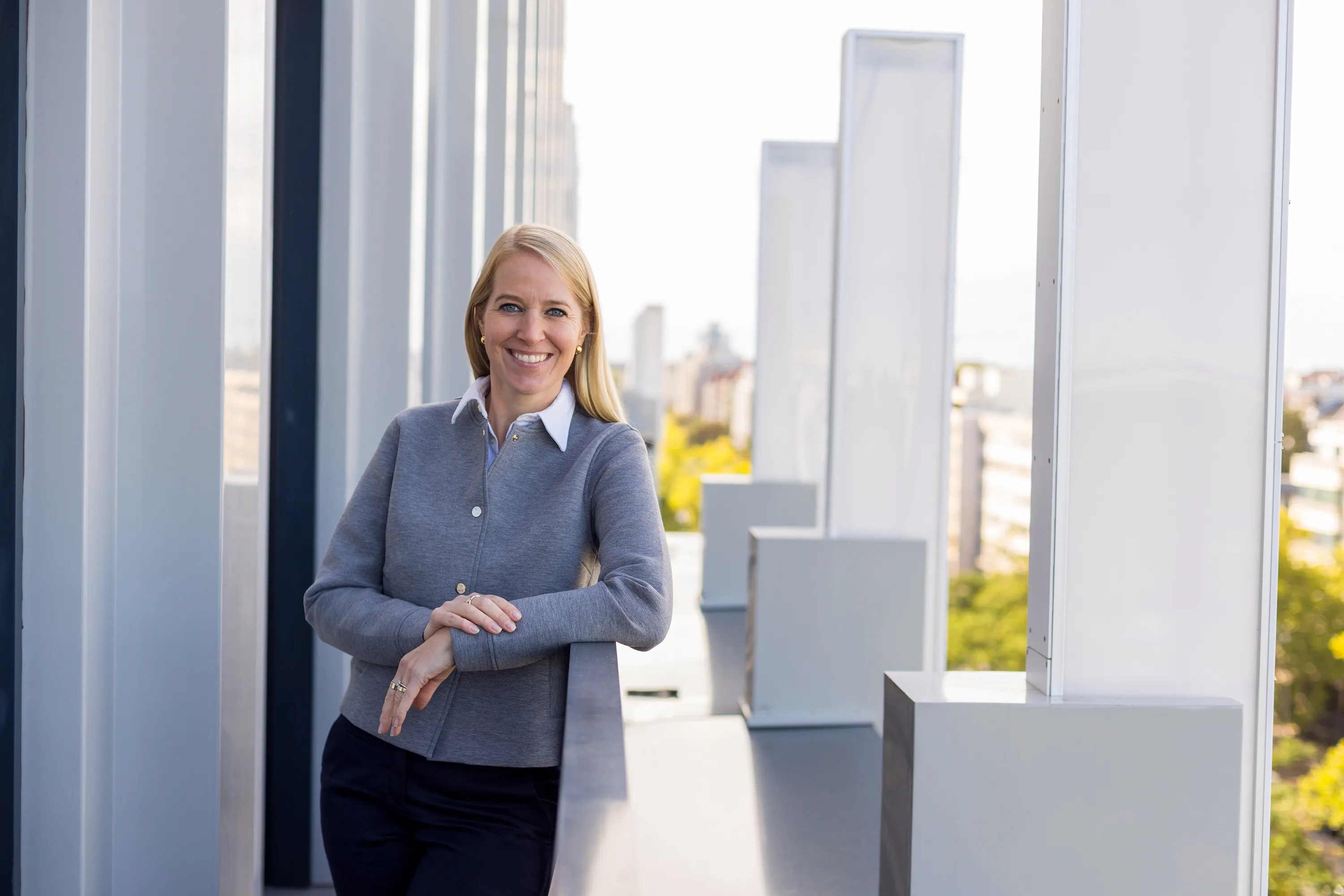 Lächelnde blonde Frau im grauen Pullover lehnt auf einem Balkon mit moderner Architektur im Hintergrund.