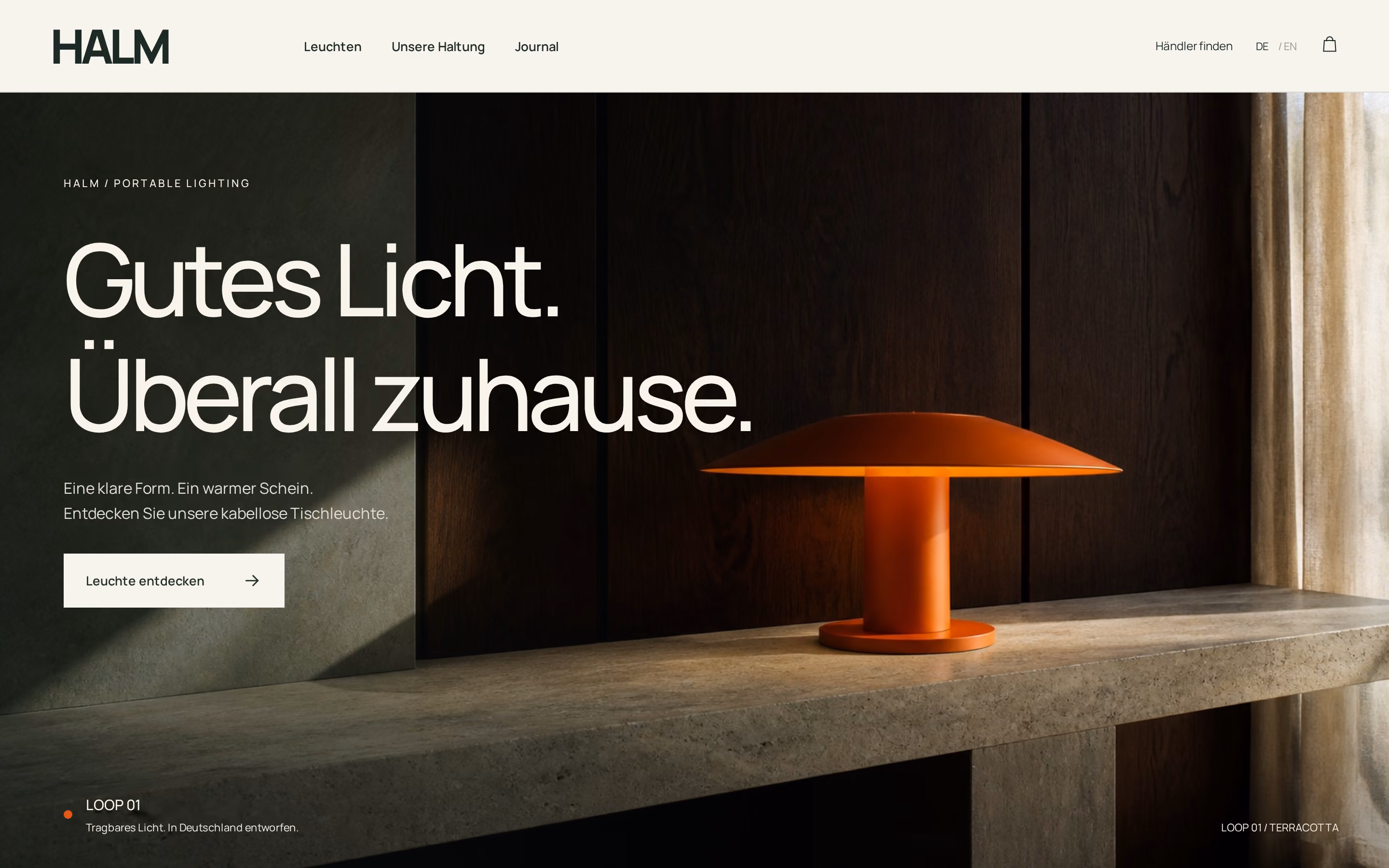 Modern orange table lamp with a wide, flat shade on a concrete shelf against a dark wooden wall with soft natural light from a window on the right.