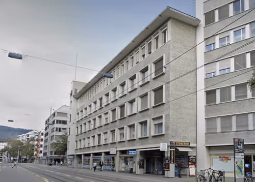 Street view of a multi-story building with shops on the ground floor and a tram line in front.