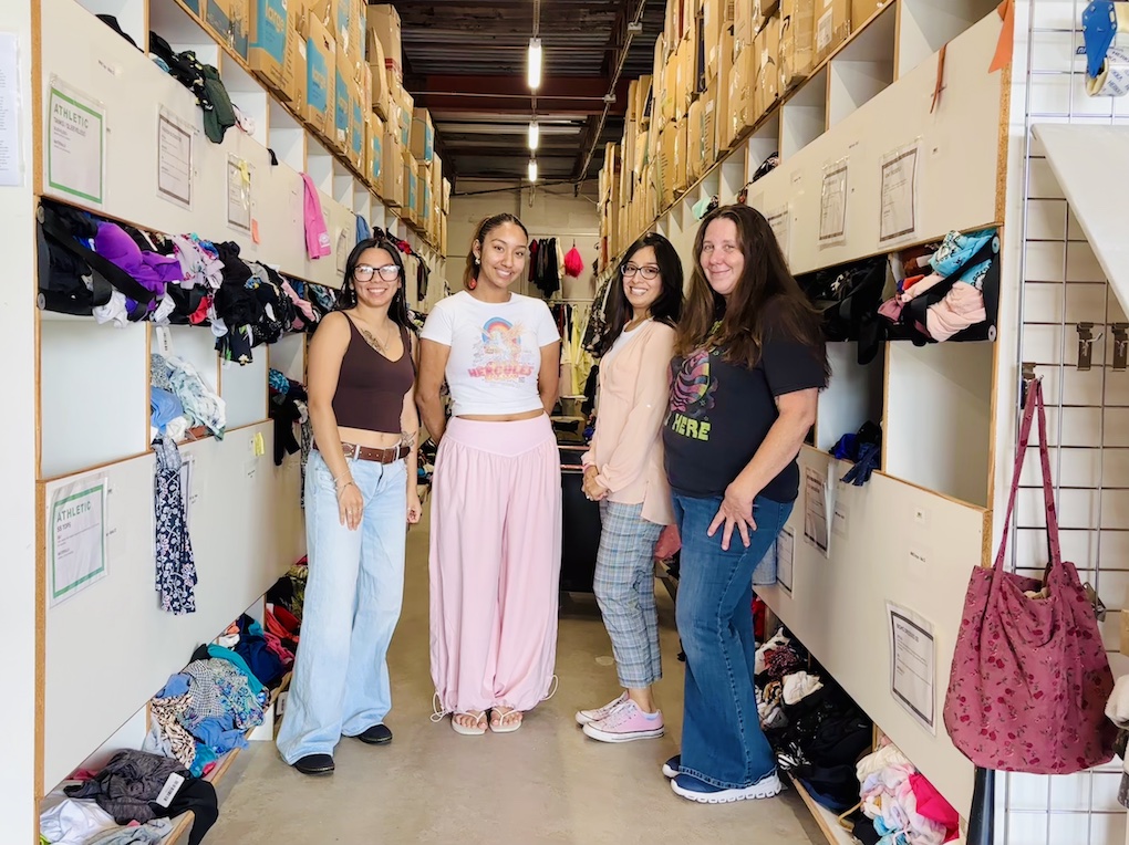 Narrow warehouse aisle with shelves on both sides holding folded clothes and labeled compartments, with hanging dresses and shoes at the far end.