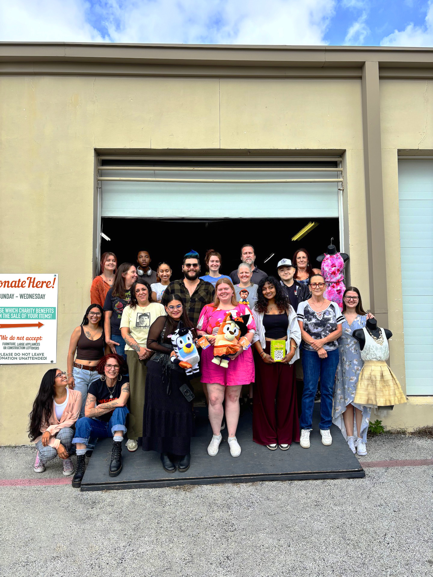 A diverse group of Thrift for Good® employees stands in front of the drive-thru donation bay at the organization’s flagship store and warehouse in Hurst, Texas, holding donated clothing, home décor, and other thrifted treasures during staff photo day. The donation bay provides a convenient drop-off location where community donations enter the warehouse to be sorted, processed, and prepared for resale, supporting local charities through sustainable shopping.