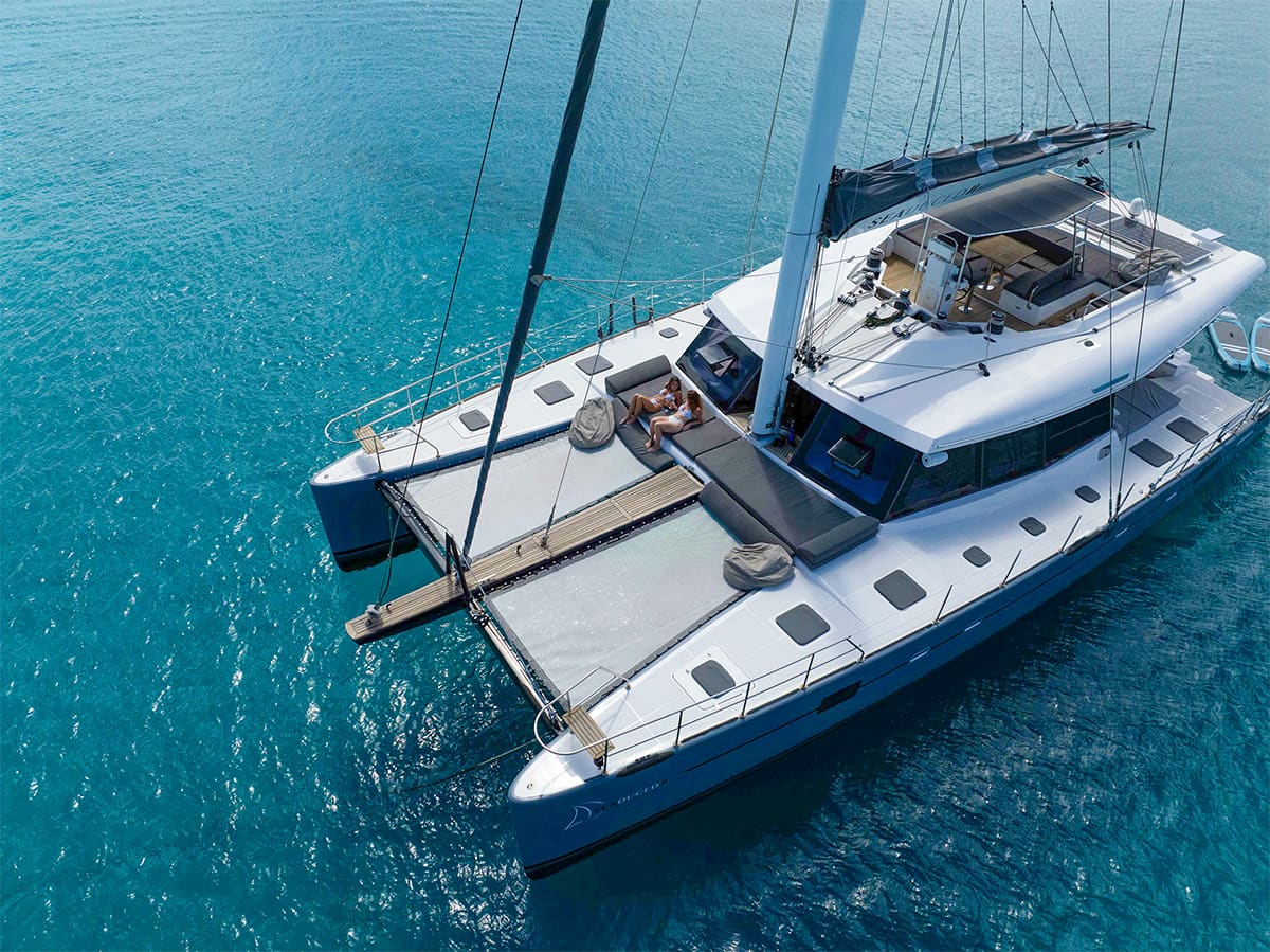 A luxurious white catamaran sailing yacht floating on clear blue water with two people relaxing on a front deck lounge.