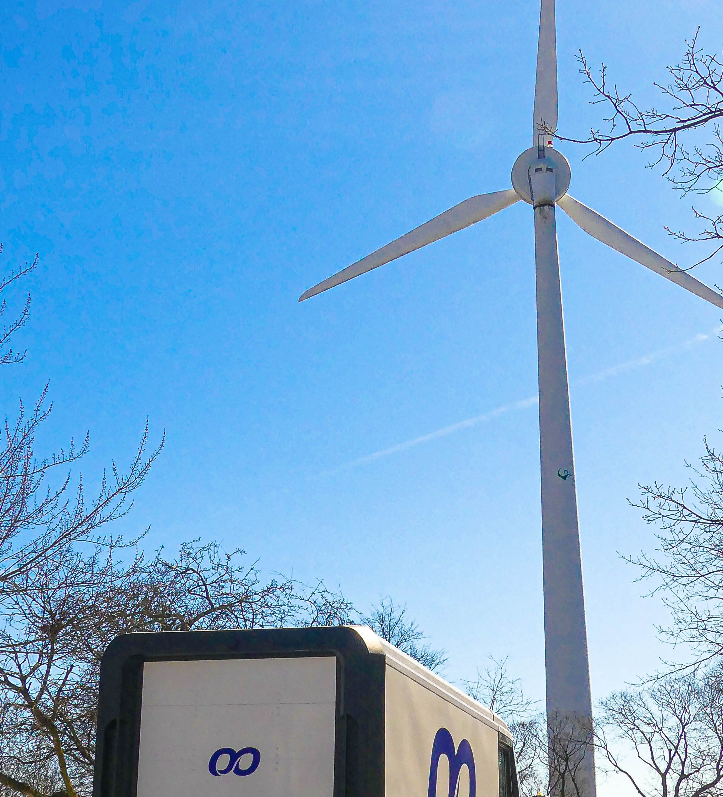 Koorier electric delivery van driving toward a large white wind turbine on a sunny day near a snowy waterfront.