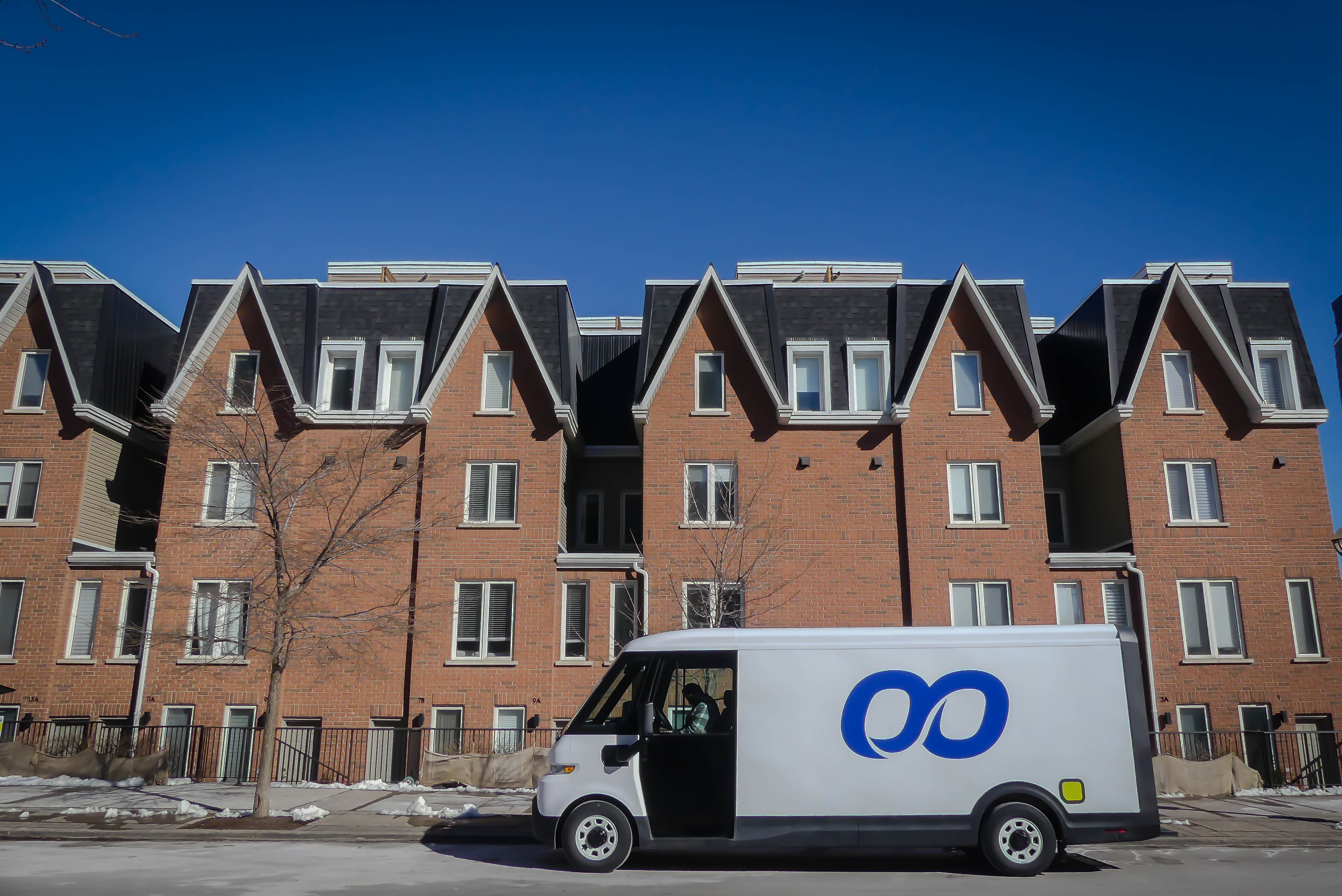 A white delivery van with a blue logo is parked on a street in front of brick townhouses under a bright blue sky, conveying a calm, urban day.