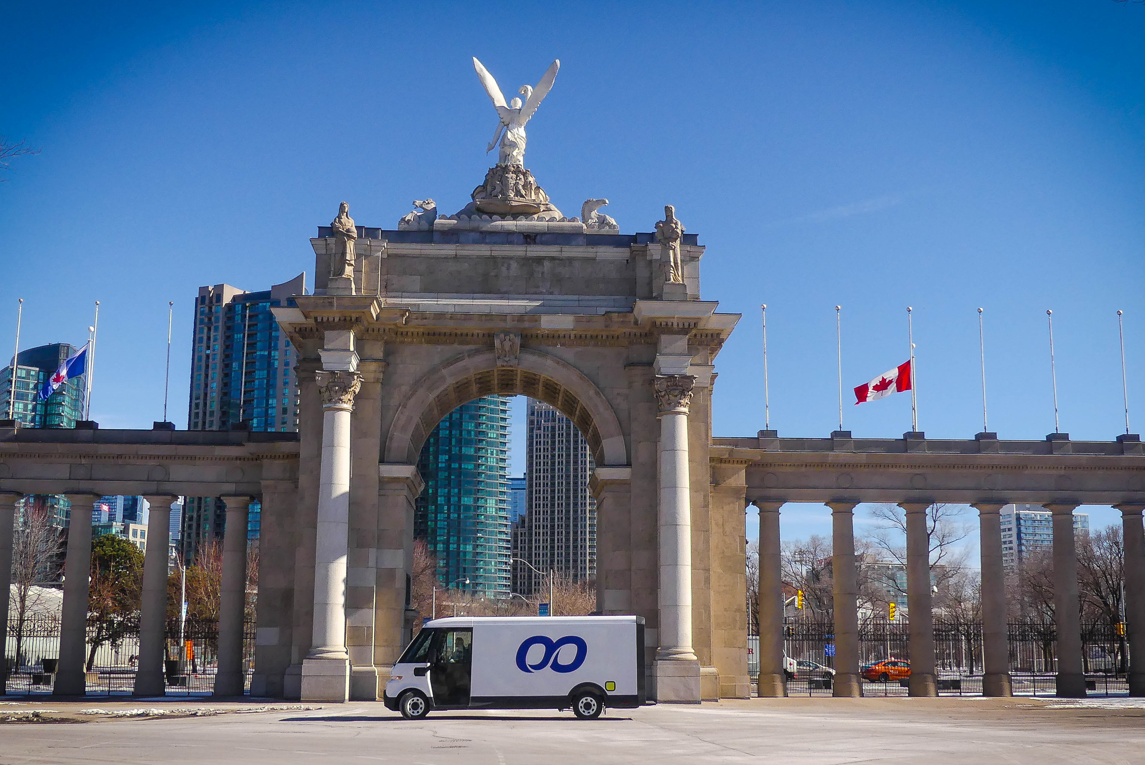 A white delivery truck with a blue infinity logo drives through a stone archway topped with a statue, against a backdrop of city buildings and a clear blue sky.