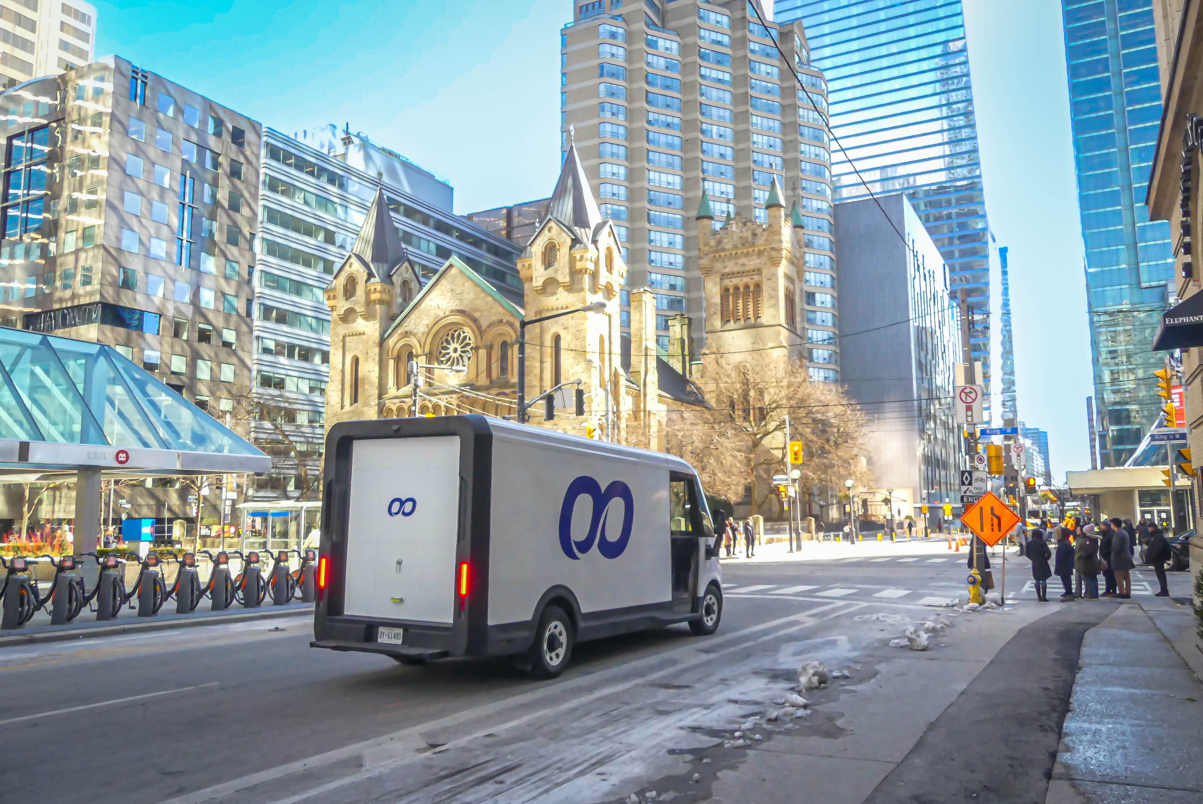 A delivery van with blue infinity logos drives through a bustling city intersection. Modern glass buildings and a historic church are illuminated by bright daylight.