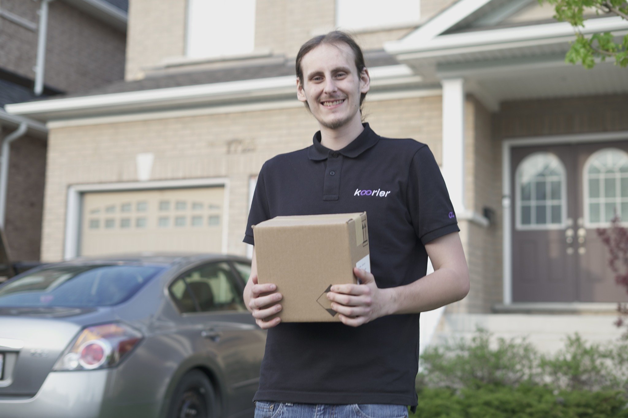 Smiling delivery person holding a package in front of a suburban house with a parked car. The person wears a black shirt. Bright, clear day.