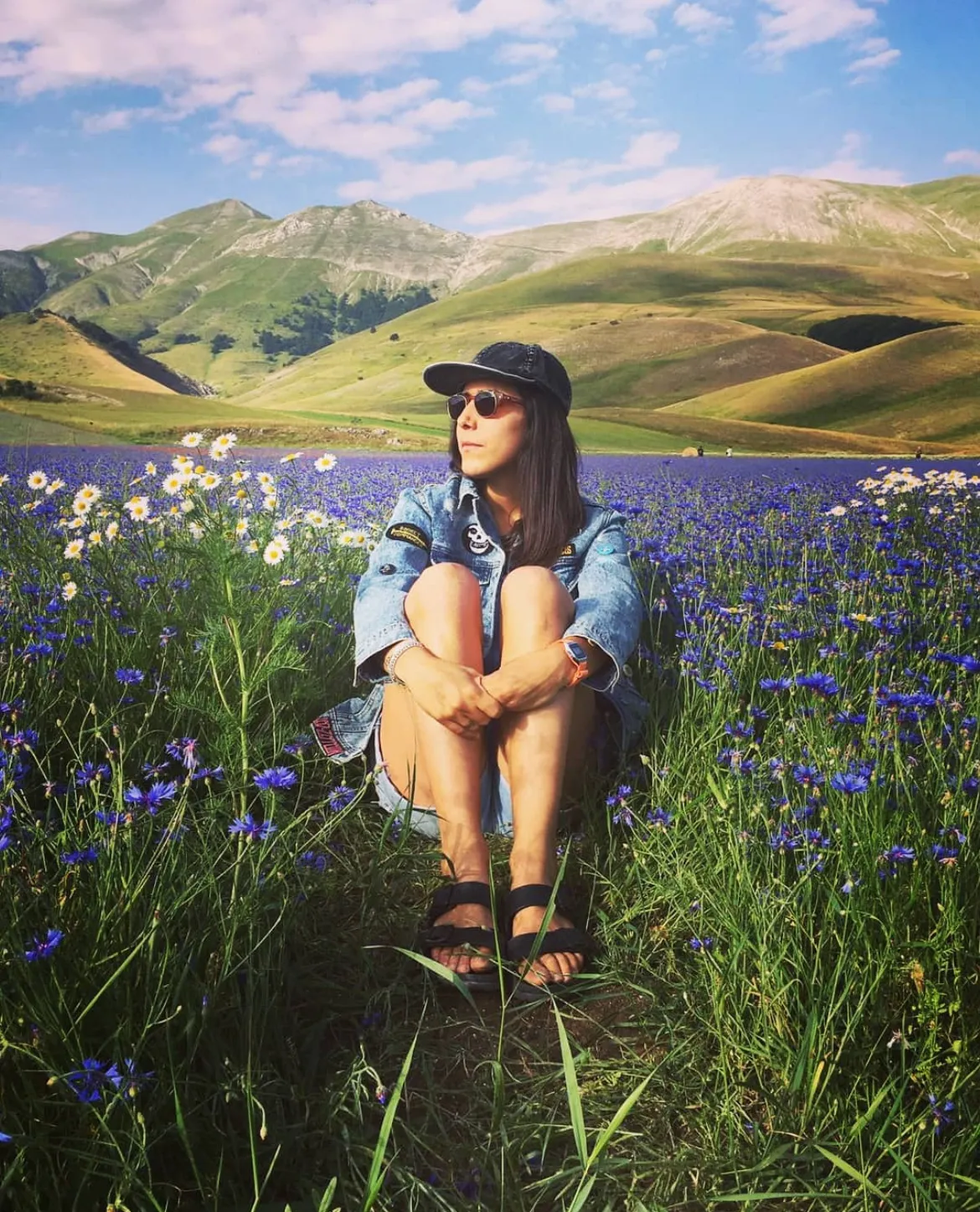 Donna seduta tra un campo di fiori viola e bianchi con colline verdi e cielo parzialmente nuvoloso sullo sfondo.