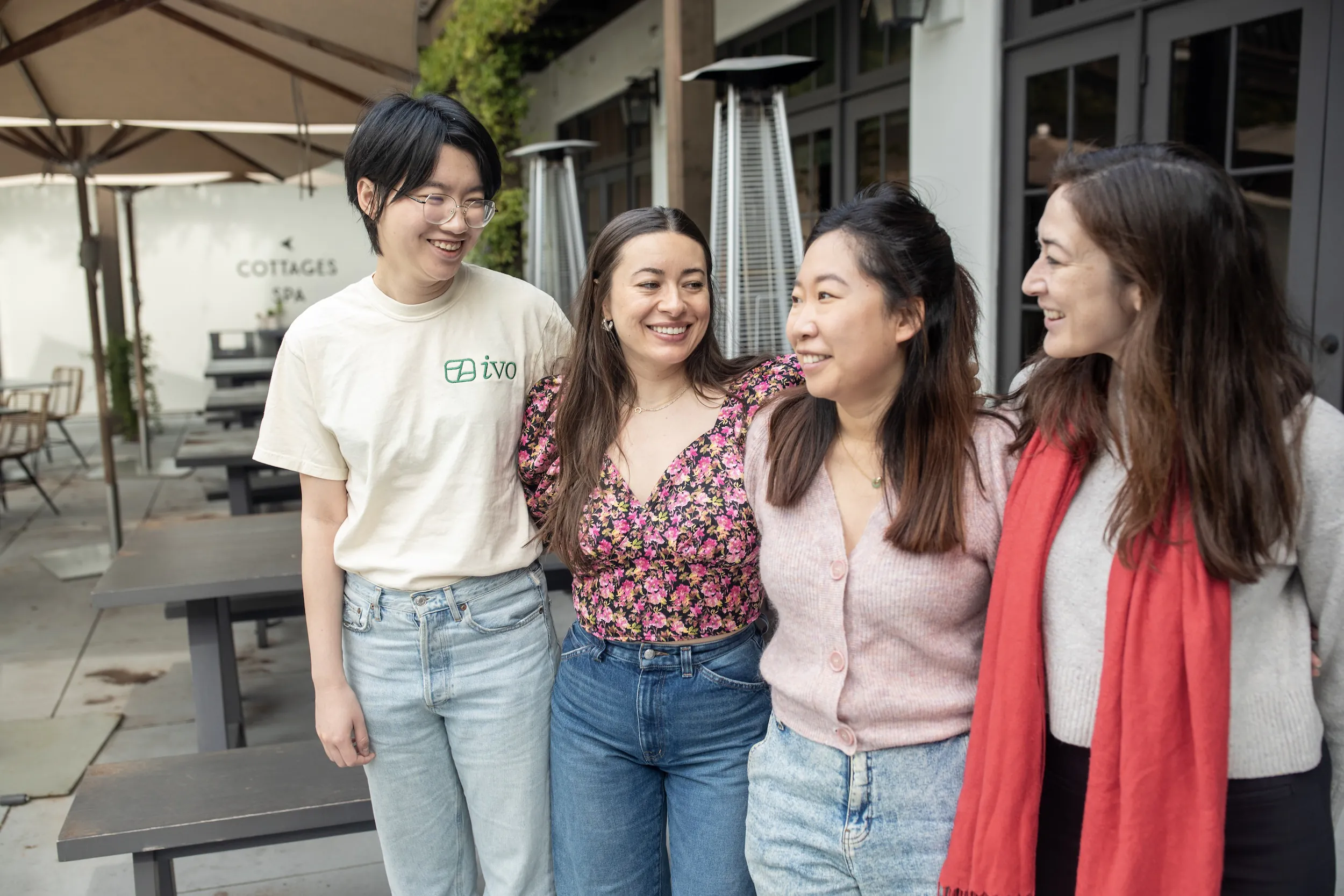 Four women standing close together, smiling and looking at each other outside a café with outdoor seating.