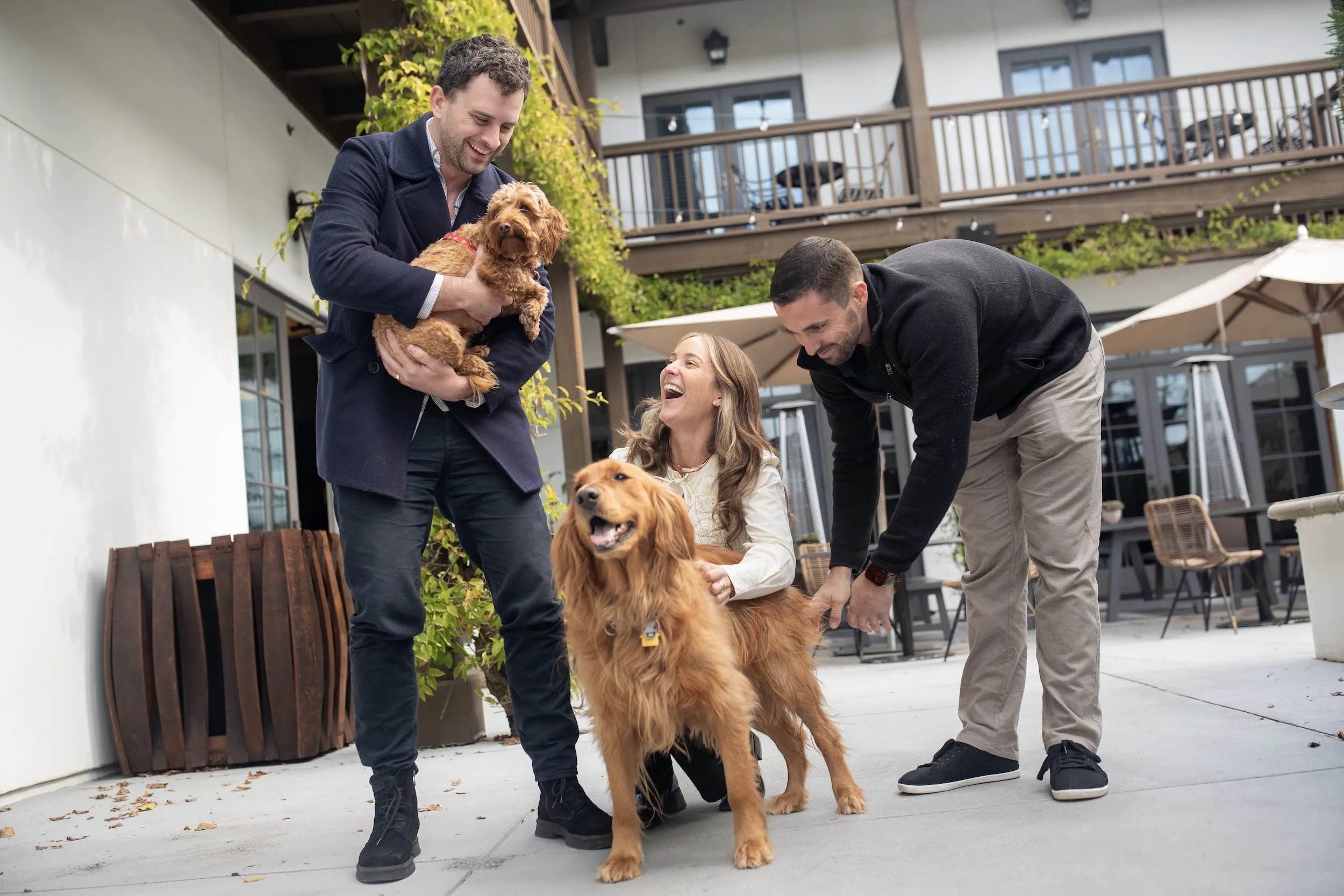 Three people joyfully interacting with a small dog being held and a golden retriever standing on a patio outside a building with balconies and outdoor seating.