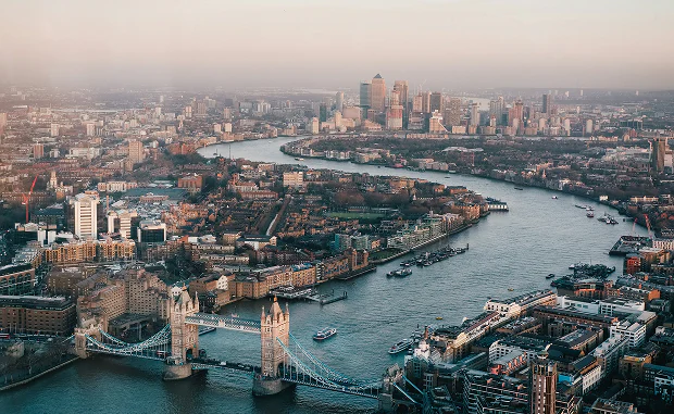 Aerial view of London's Tower Bridge spanning the River Thames with city buildings and Canary Wharf skyline in the background.