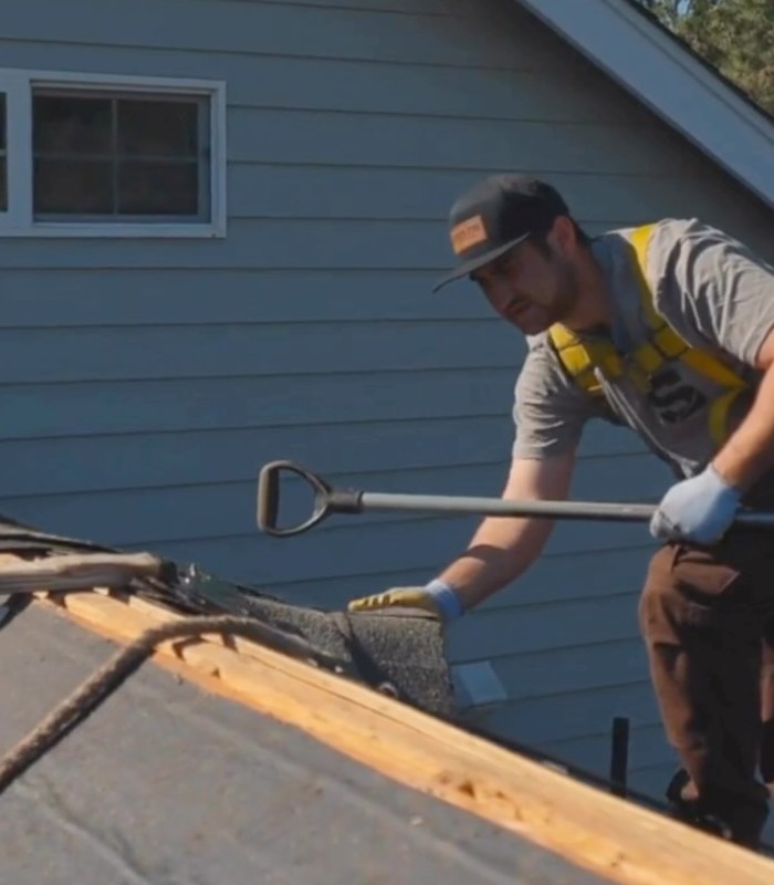 Worker wearing safety harness installs or repairs roofing shingles using a tool on a house roof.