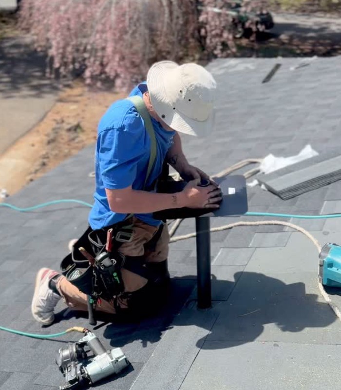 Roofer kneeling on shingled roof installing or repairing a pipe vent with tools nearby and safety harness.