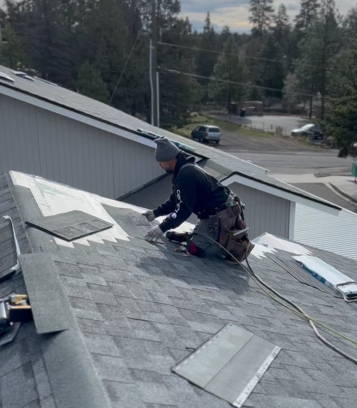 Worker wearing gloves and a beanie installing asphalt shingles on a rooftop during daytime.