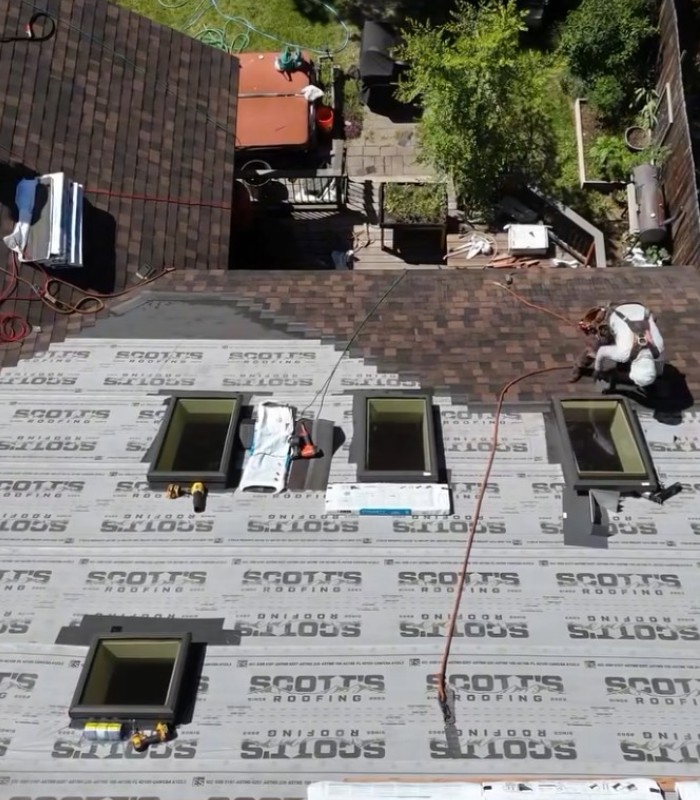 Worker installs shingles on a roof with four skylights and underlayment labeled Scott's Roofing.
