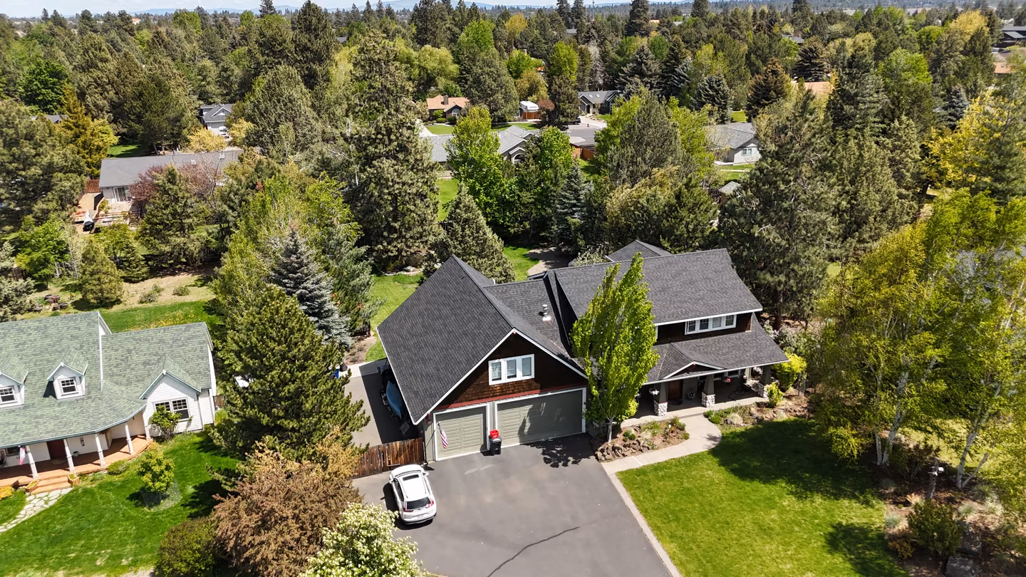 Aerial view of a suburban neighborhood with a large house featuring a dark gray roof, two-car garage, and driveway with a white vehicle parked. The property is surrounded by green lawns and numerous tall trees, with other houses and dense woods visible in the background under a clear sky.