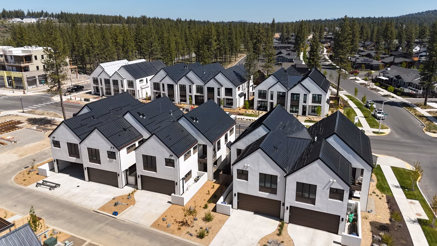 Aerial view of a modern residential neighborhood with rows of white two-story houses featuring dark roofs, garages, paved driveways, and landscaping, surrounded by trees.