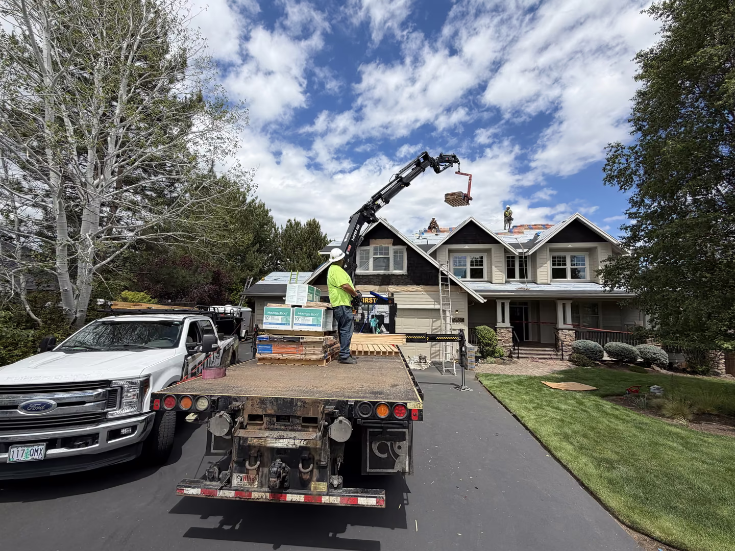 Construction workers on roof of a two-story house with a crane lifting roofing materials from a flatbed truck parked in driveway beside a white pickup truck, under a partly cloudy sky.