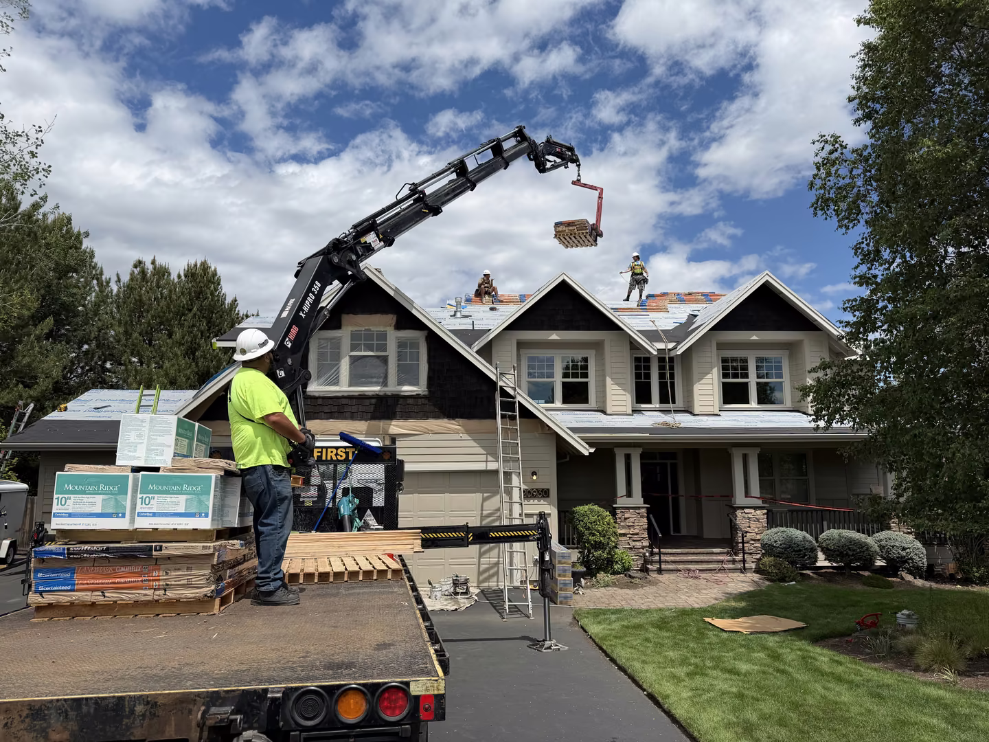 Construction workers on roof of two-story house using a crane on a truck to lift roofing materials while another worker controls the crane from the truck bed, under a partly cloudy sky.