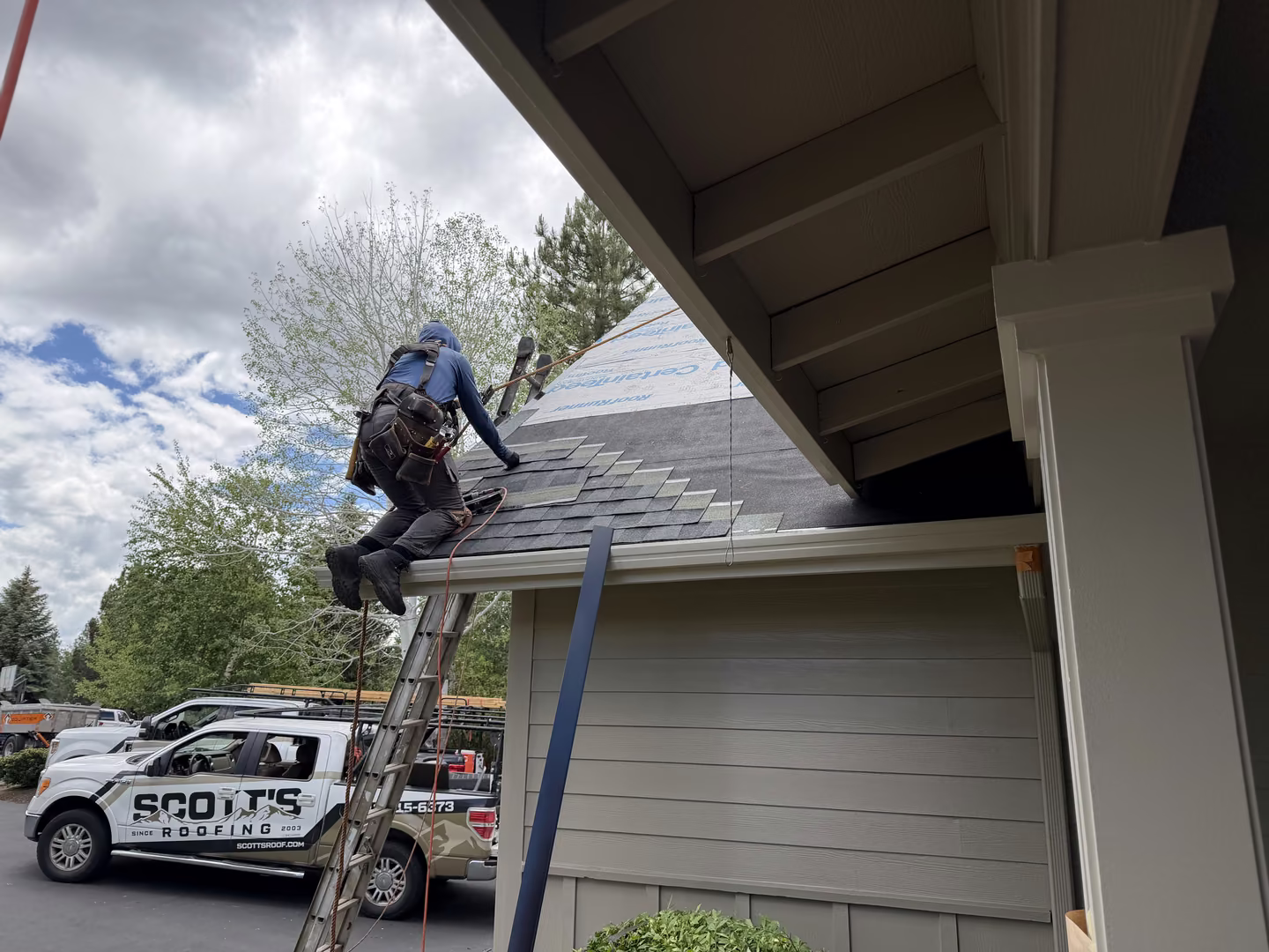 Roofer wearing safety gear climbing a ladder to install shingles on a house roof with a company truck labeled 'Scott's Roofing' parked nearby under a cloudy sky.