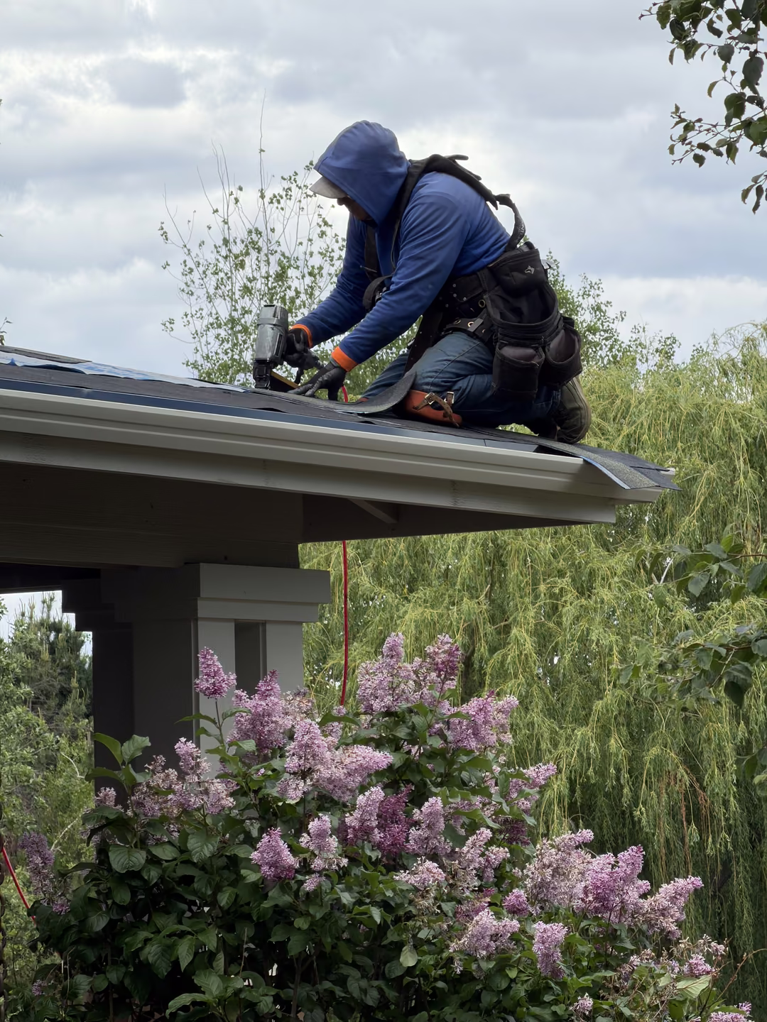 A worker wearing a blue hoodie and safety harness is kneeling on a roof edge using a nail gun for roofing work, surrounded by green trees and blooming lilac flowers below.