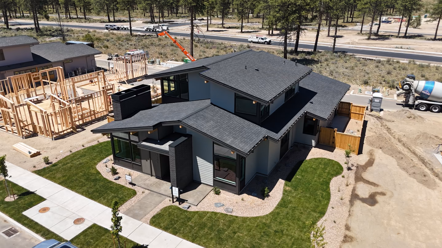 A modern two-story house with a dark roof and gray exterior surrounded by a green lawn and landscaping, next to a construction site with wooden framing and a cement mixer truck.