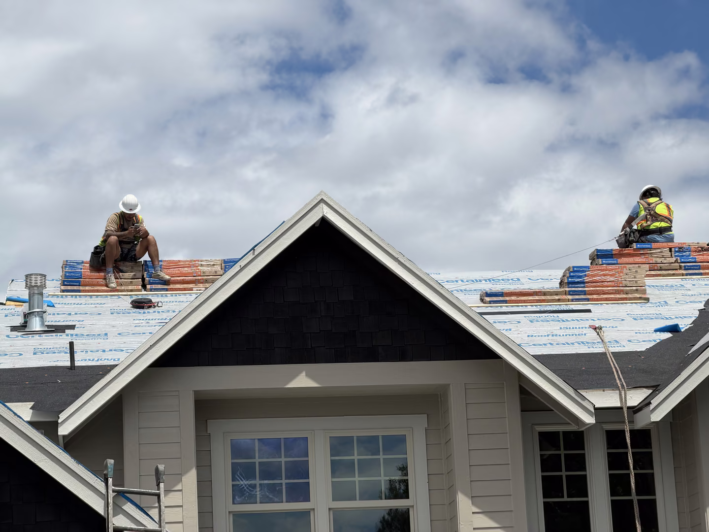 Two construction workers wearing helmets and safety harnesses installing roofing materials on the sloped roof of a beige house under a partly cloudy blue sky.