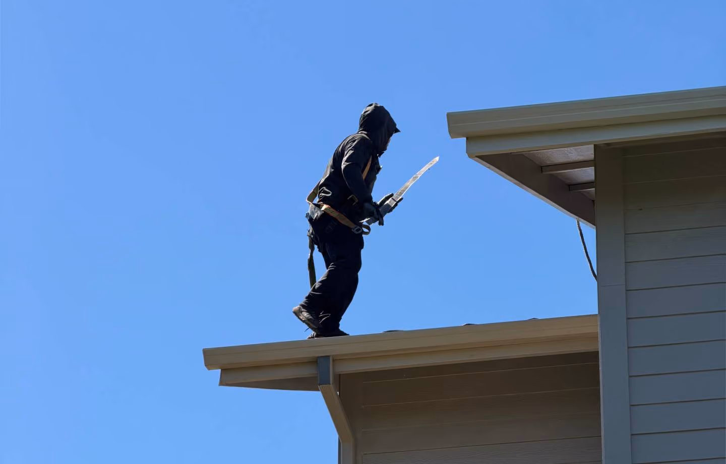 Person wearing black clothing and a hood using a safety harness stands on the edge of a roof holding a tool against a clear blue sky.